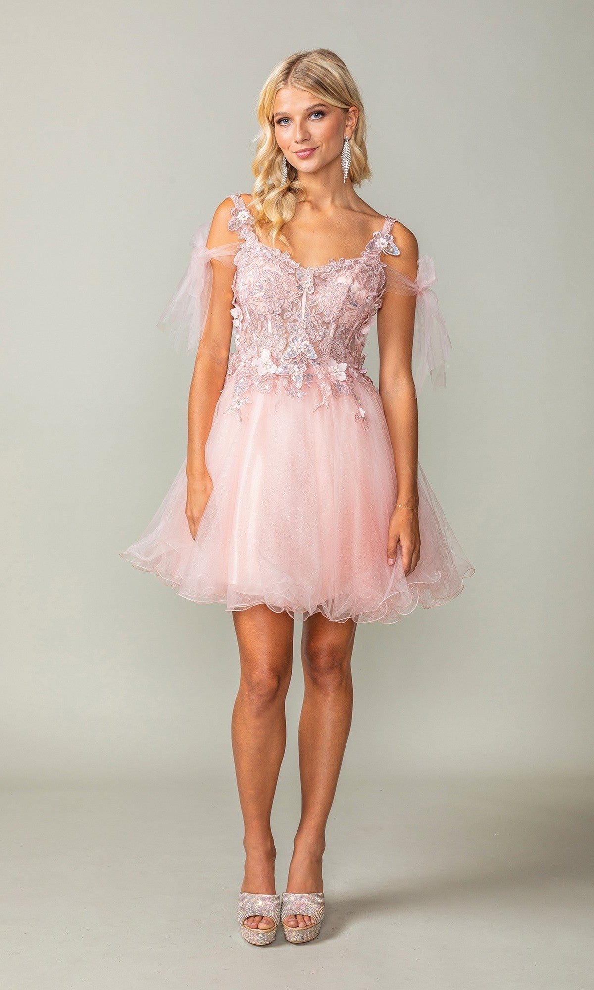 A woman stands against a plain light background wearing the Dancing Queen 3365 short homecoming dress in blush, featuring an off-shoulder neckline, lace details, and a glitter tulle skirt.